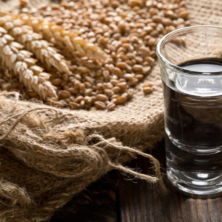 A glass of clear schnapps stands next to ears of grain on a coarse jute sack.