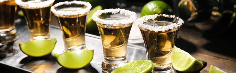 Fünf Tequila-Shots mit Salzrand stehen neben Limettenscheiben auf einem Tablett in dunkler Umgebung.
