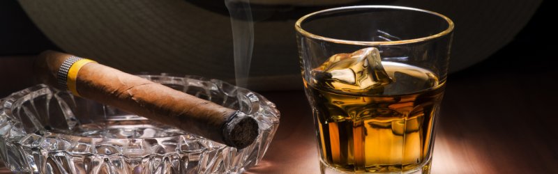 Zigarre raucht in Aschenbecher neben Whiskyglas mit Eis auf Holztisch im schummrigen Licht.