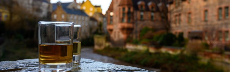 Two whisky glasses stand on a wall, with blurred buildings in the background.