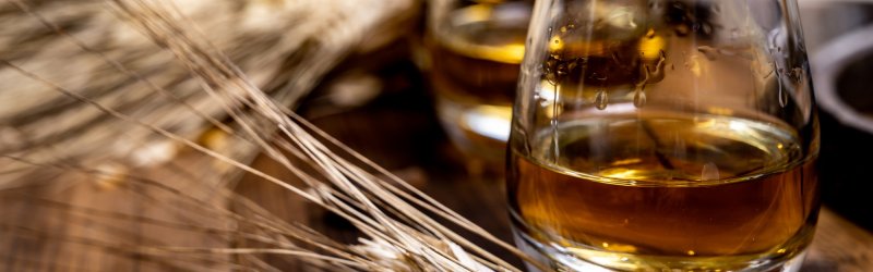 Ein Glas Whisky steht auf einem Holztisch neben trockenen Weizenähren.