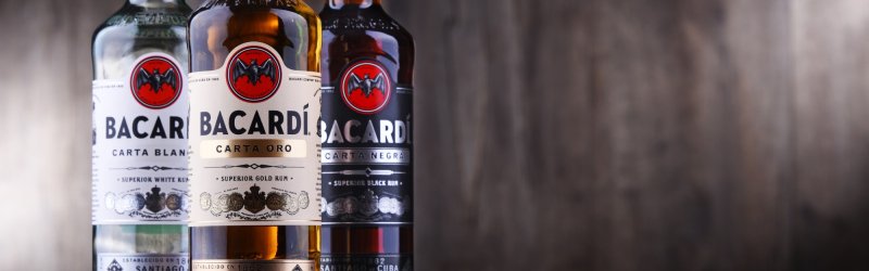 Three bottles of rum stand next to each other against a blurred, wooden background: Bacardi Carta Blanca, Oro, Negra.