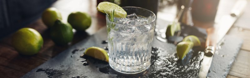 A glass filled with ice and decorated with limes stands on a wet wooden table.