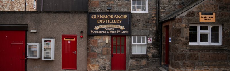 Stone house with red door, labeled "Glenmorangie Distillery", on a cobbled square under a cloudy sky.