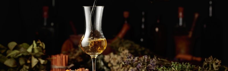 Goldene Flüssigkeit wird in ein Glas gegossen; umgeben von Kräutern und Gewürzen.