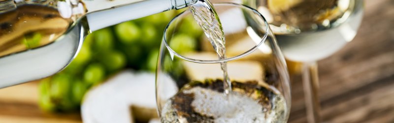 White wine is poured into a glass, accompanied by grapes and cheese on a wooden table.