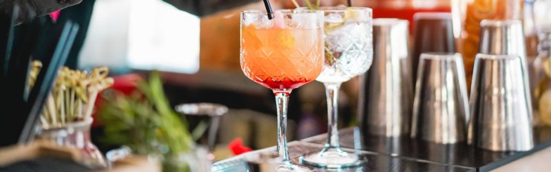 Glasses - standing - on a bar with various cocktails and metallic shakers in the background.