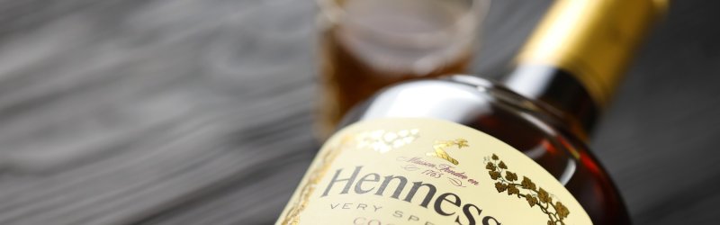 Flasche Hennessy Cognac liegt auf Tisch, Glas im Hintergrund, schwarz-grauer Holzuntergrund.