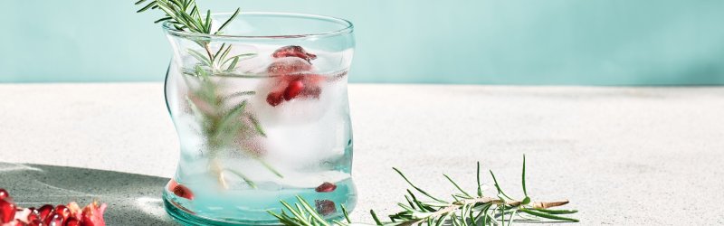 Glass with rosemary and pomegranate seeds stands on a table against a light blue background.