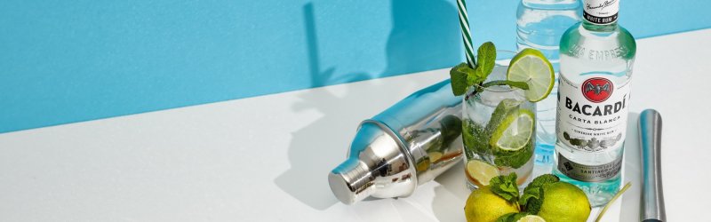 Les ingrédients du cocktail sont disposés sur la table ; bouteille de Bacardi, citrons verts, thé glacé, shaker, menthe, fond bleu et blanc.