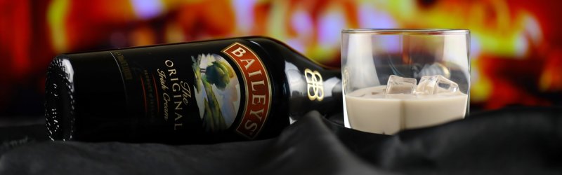 Fles Baileys ligt naast glas met ijsblokjes op zwart doek, kleurrijke achtergrondverlichting.
