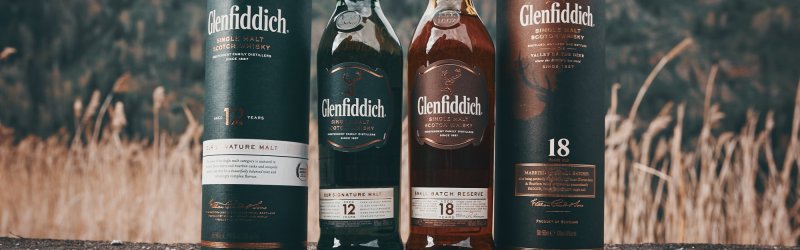De flessen en blikjes van Glenfiddich staan op een stenige ondergrond tegen een achtergrond van beboste heuvels.