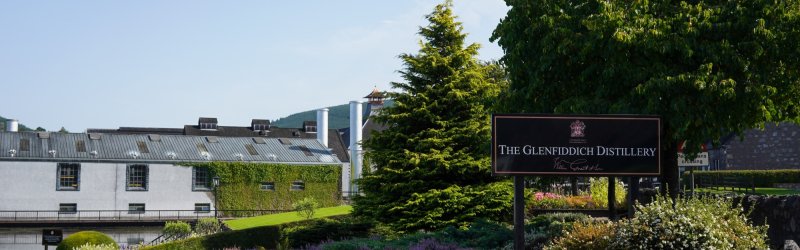 Het bord van de Glenfiddich distilleerderij staat in de groene tuin voor de industriële gebouwen.