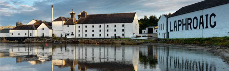 Weiße Gebäude spiegeln sich im ruhigen Wasser; Aufschrift: „LAPHROAIG“. Bewölkter Himmel im Hintergrund.