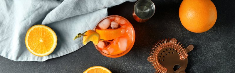 Ein Cocktailglas mit Eis steht neben Orangenhälften, einem Messbecher und einem Bar-Sieb.