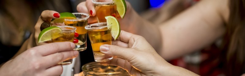 Hands toast with tequila shots, garnished with limes, in a bar-like setting.