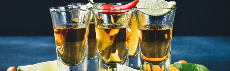 Four tequila glasses stand on a black surface, surrounded by limes, nuts and salt.