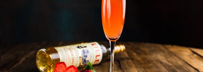 A cocktail glass with a red drink stands on a wooden table, next to strawberries and a horizontal bottle.
