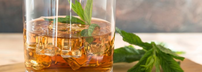 Glas mit Whisky und Eiswürfeln, dekoriert mit Minzblättern, steht auf einem Holzuntergrund.