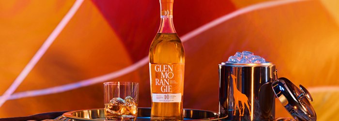 Bottle of whisky with glass and ice bucket on tray, in front of colorful, patterned background.