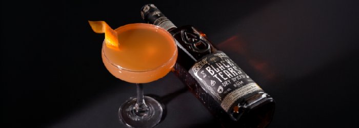 Een oranje cocktail in een glas naast een Black Tears fles met een zwarte achtergrond