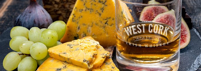 A bird's eye view shows a rustic wooden board with cheese, grapes, figs and a glass of amber liquid against a blurred background. The scene is well lit, making the textures and colors of the e