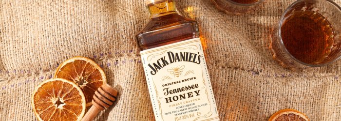 Fles Jack Daniel's Tennessee Honey ligt op jute doek, omringd door glazen, kaneelstokjes en sinaasappelschijfjes.