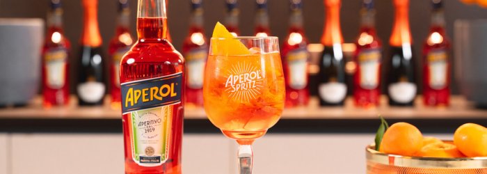 Aperol bottle and Aperol spritz glass on the table, oranges and bottles in the background.