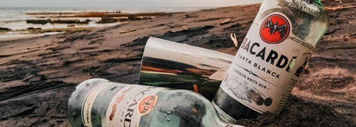 Deux bouteilles de Bacardi sont posées sur le sable d'une plage, ciel nuageux, mer en arrière-plan.