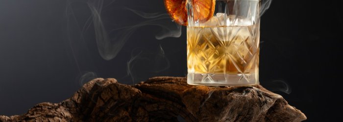 Whiskey glass with orange slice stands smoky on a rustic piece of wood in a dark background.