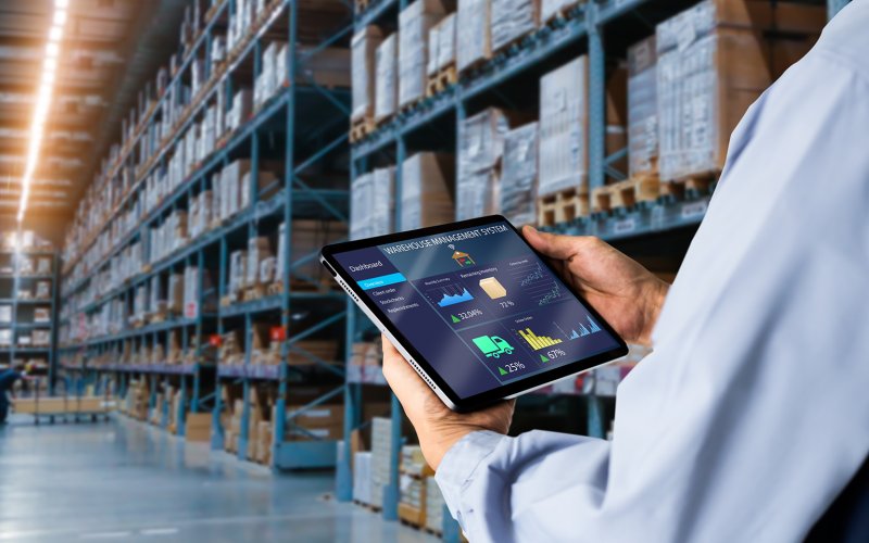 Tablet toont magazijnbeheer, vastgehouden door persoon in blauw shirt, voor goed gevulde magazijnrekken.