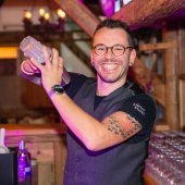 A bartender smiles and shakes a cocktail shaker in a cozy bar setting.