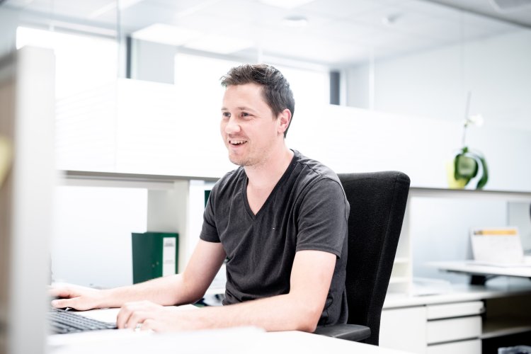Een man zit glimlachend aan zijn bureau, werkend aan zijn computer in een modern kantoor.